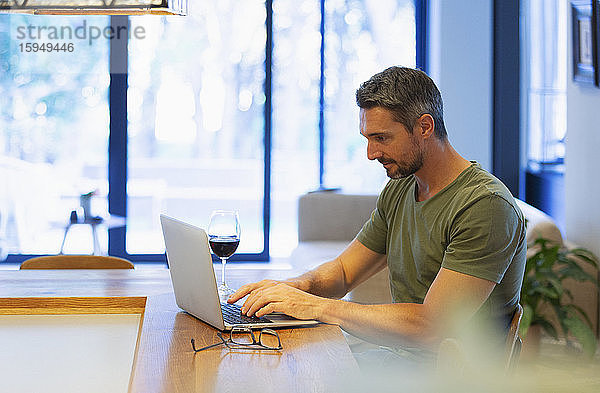 Mann benutzt Laptop und trinkt Wein  arbeitet von zu Hause in der Küche