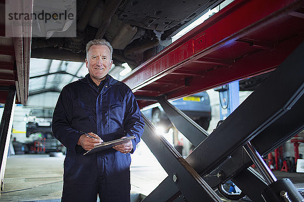 Porträt eines selbstbewussten  lächelnden männlichen Mechanikers mit Klemmbrett  der in einer Autowerkstatt unter dem Auto arbeitet
