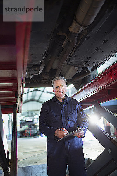 Porträt eines selbstbewussten männlichen Mechanikers mit Klemmbrett  der in einer Autowerkstatt unter dem Auto arbeitet