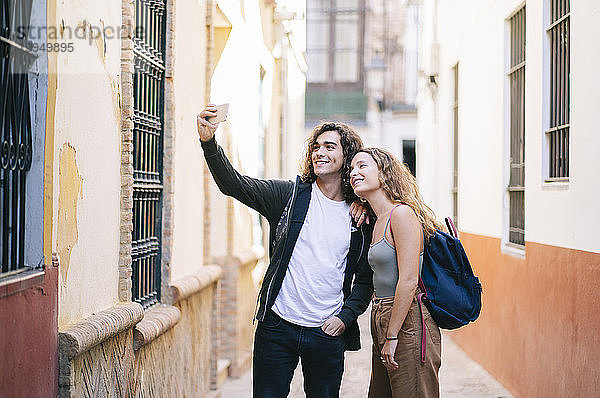 Lächelndes junges Paar  das sich beim Stehen in einer engen Straße in Santa Cruz  Sevilla  Spanien  selbstständig macht