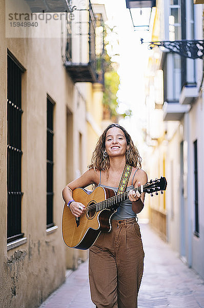 Porträt einer lächelnden jungen Frau  die Gitarre spielt  während sie auf einer schmalen Straße in Santa Cruz  Sevilla  Spanien  spazieren geht