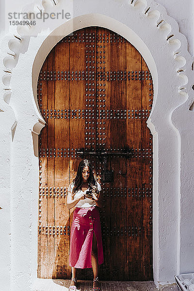 Junge Frau steht vor der traditionellen Eingangstür und schaut auf Handy  Merzouga  Marokko