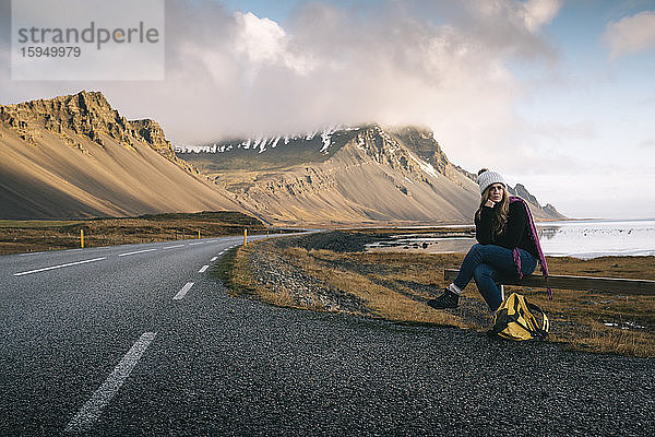 Frau mit Rucksack sitzt auf einem Geländer  während sie am Straßenrand gegen die Berge in Island wartet
