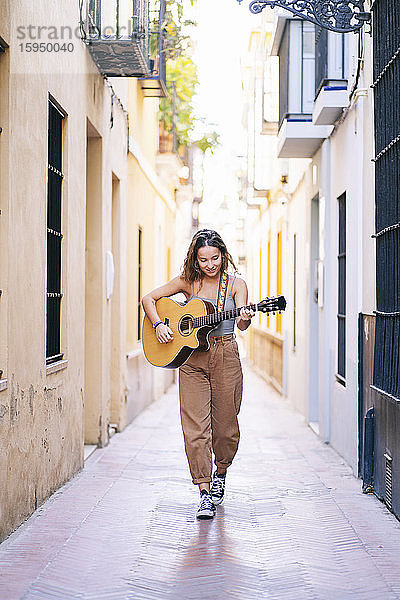 Junge Frau in voller Länge spielt Gitarre  während sie auf einer schmalen Straße inmitten von Gebäuden in Santa Cruz  Sevilla  Spanien