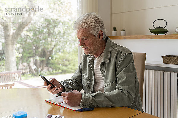 Älterer diabetischer Mann benutzt Mobiltelefon  während er zu Hause in sein Tagebuch schreibt