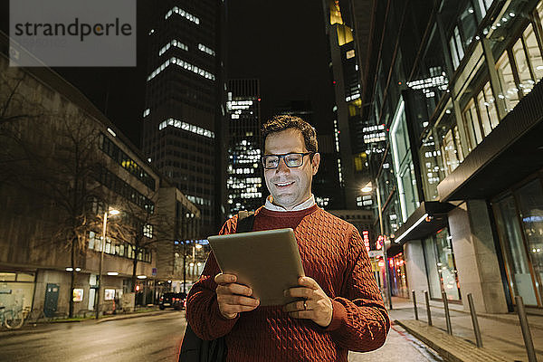 Portrait eines lächelnden Unternehmers in der Stadt  der nachts auf ein digitales Tablet schaut  Frankfurt  Deutschland