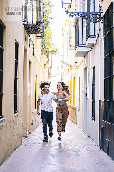Ein glückliches junges Paar läuft in voller Länge auf einer schmalen Straße in Santa Cruz  Sevilla  Spanien