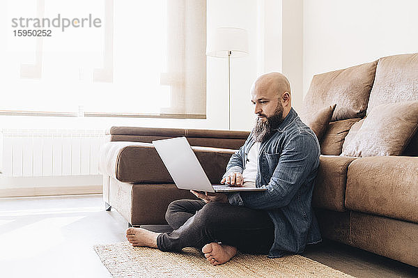Geschäftsmann mit rasiertem Kopf  der am Laptop arbeitet  während er zu Hause am Boden vor dem Sofa sitzt