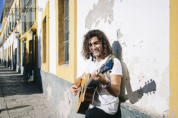 Lächelnder junger Mann spielt Gitarre  während er an der Wand lehnt  Santa Cruz  Sevilla  Spanien