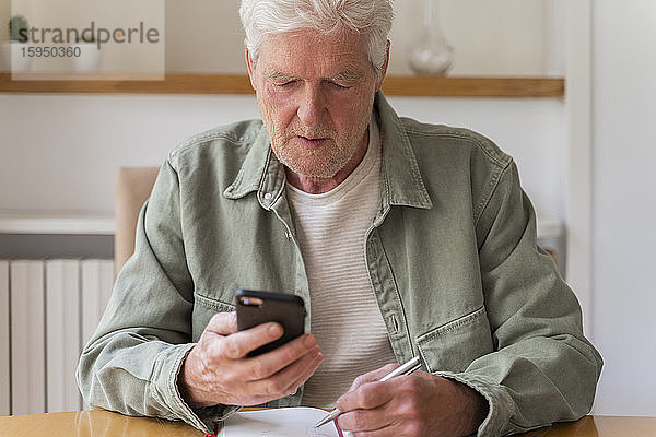 Pensionierter zuckerkranker älterer Mann benutzt Smartphone  während er zu Hause mit seinem Tagebuch sitzt
