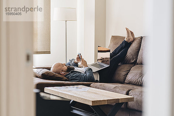 Geschäftsmann mit Laptop  der zu Hause auf dem Sofa liegend über sein Smartphone Musik hört