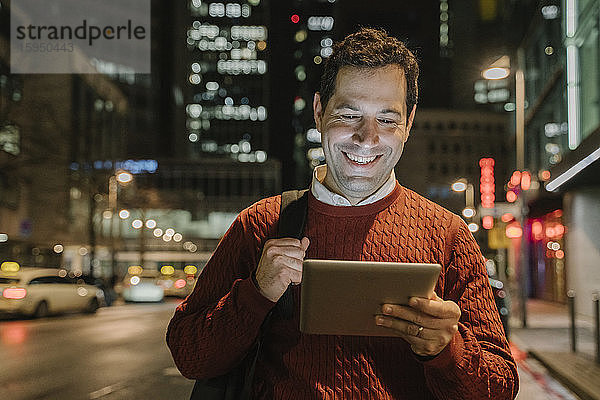 Portrait eines glücklichen Unternehmers in der Stadt  der nachts auf ein digitales Tablet schaut  Frankfurt  Deutschland