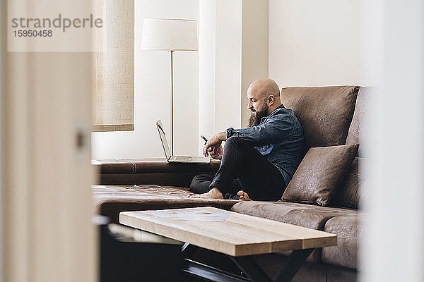 Geschäftsmann mit Laptop  der sein Handy benutzt  während er zu Hause auf dem Sofa sitzt