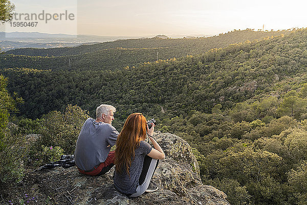 Vater und erwachsene Tochter sitzen auf einem Felsen und genießen die Aussicht bei Sonnenuntergang