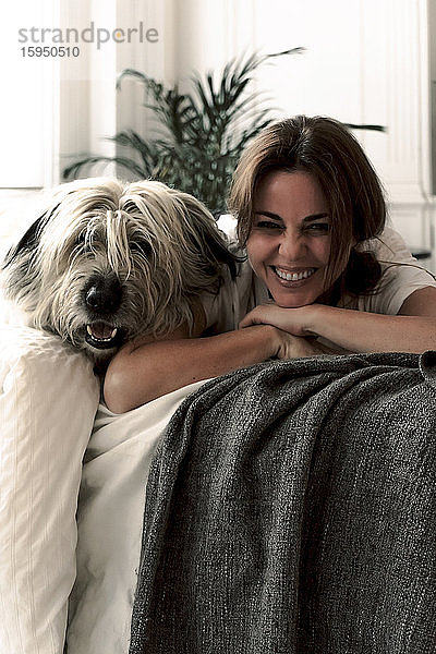 Porträt einer lachenden  reifen Frau  die mit ihrem Hund auf dem Bett liegt