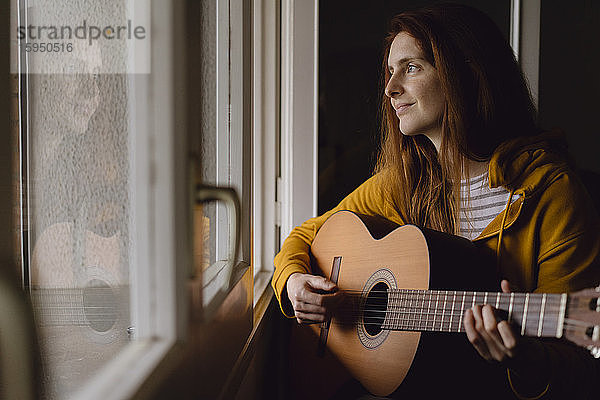 Porträt einer entspannten rothaarigen Gitarre spielenden Frau am offenen Fenster