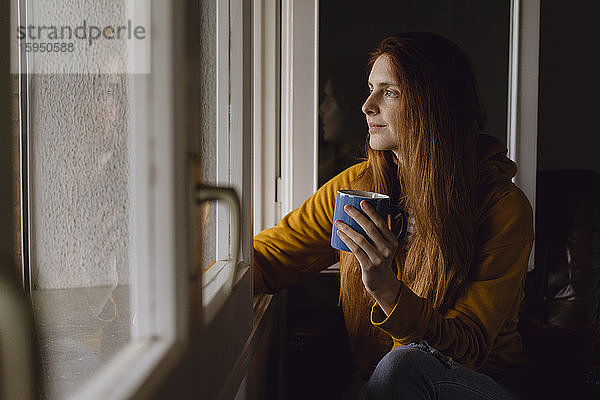 Lächelnde rothaarige Frau mit Kaffeetasse  die aus dem offenen Fenster schaut