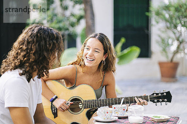 Glückliche junge Frau spielt Gitarre  während sie mit ihrem Freund in einem Straßencafé sitzt  Santa Cruz  Sevilla  Spanien