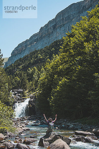 Spanien  Provinz Huesca  Wanderin hebt die Arme und bewundert kleinen Wasserfall am klaren Bergfluss