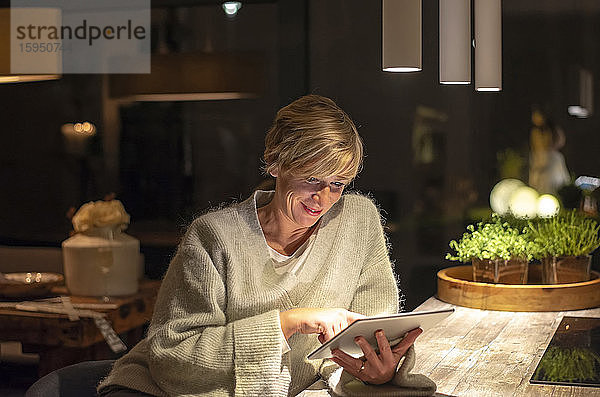 Lächelnde blonde Frau arbeitet spät  während sie ein digitales Tablet im beleuchteten Wohnzimmer benutzt