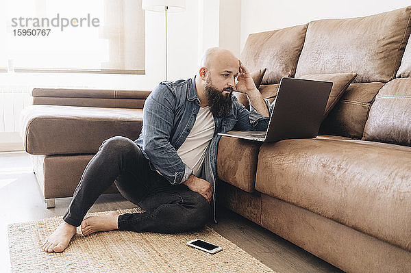 Seriöser Geschäftsmann mit Laptop  während er zu Hause auf dem Boden am Sofa sitzt