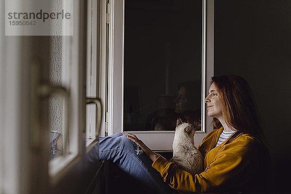 Lächelnde rothaarige Frau sitzt mit ihrer Katze am offenen Fenster und schaut in die Ferne