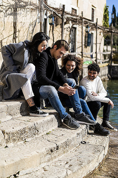 Vier Freunde sitzen auf einer Treppe am Gardasee und teilen sich ein Smartphone  Italien