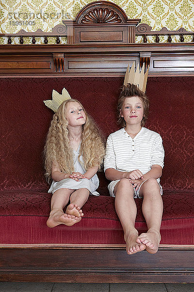 Junge und Mädchen sitzen auf einer Couch und tragen Pappkronen