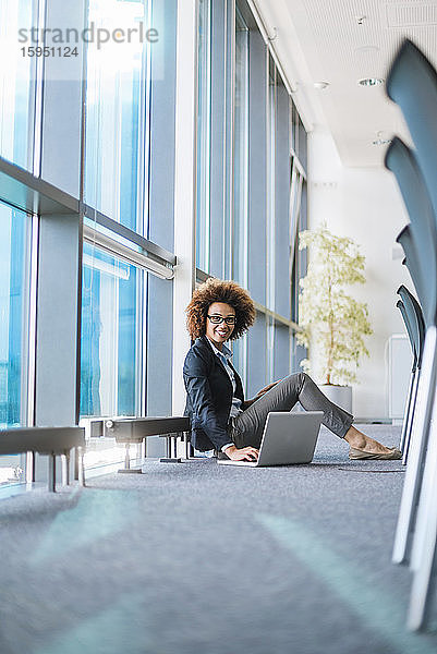 Lächelnde junge Geschäftsfrau sitzt mit Laptop auf dem Boden im Konferenzraum