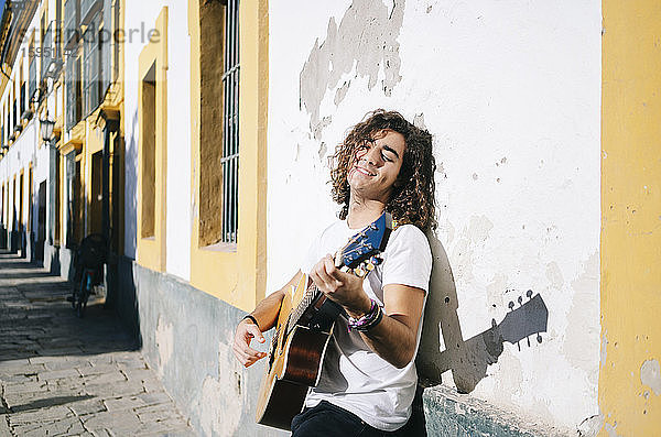 Lächelnder junger Mann mit geschlossenen Augen spielt Gitarre  während er an einer Wand lehnt  in Santa Cruz  Sevilla  Spanien