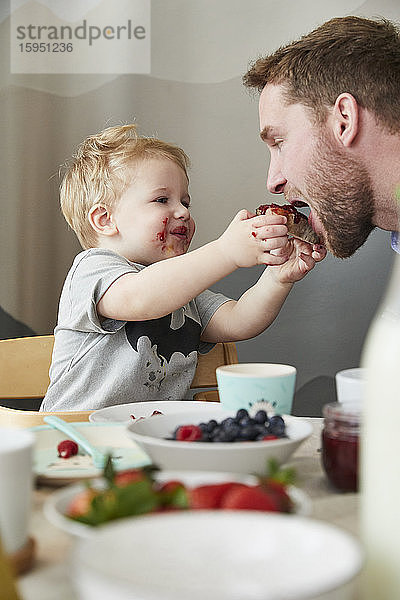 Porträt eines verschmierten kleinen Jungen  der seinen Vater am Frühstückstisch mit Marmeladenbrot füttert