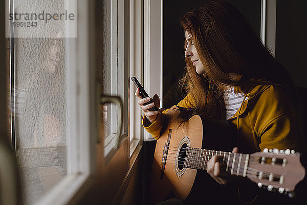 Lächelnde rothaarige Frau mit Gitarre sitzt am offenen Fenster und schaut auf ihr Handy