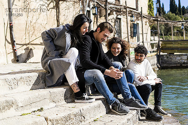 Vier Freunde sitzen auf einer Treppe am Gardasee und teilen sich ein Smartphone  Italien