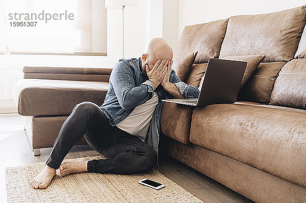 Müde Geschäftsmann mit Laptop bedeckt Gesicht  während er zu Hause im Wohnzimmer sitzt