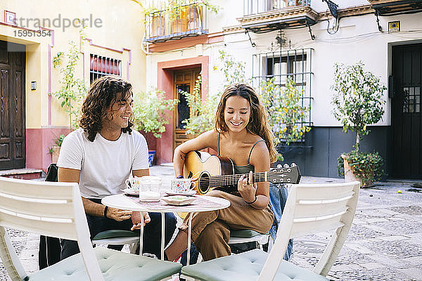 Lächelnder junger Mann sieht seine Gitarre spielende Freundin an  während er in einem Straßencafé sitzt  Santa Cruz  Sevilla  Spanien