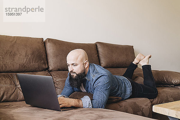 Geschäftsmann benutzt Laptop  während er zu Hause auf dem Sofa im Wohnzimmer liegt