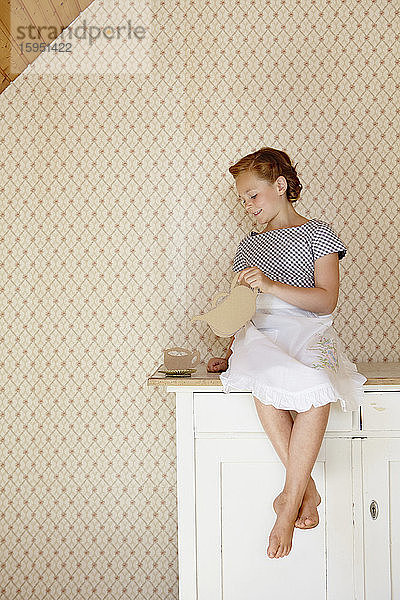 Mädchen mit Schürze spielt mit einem Teeservice aus Pappe auf der Kommode