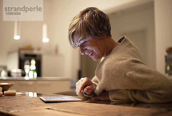 Lächelnde Frau arbeitet spät  während sie ein digitales Tablet im beleuchteten Wohnzimmer benutzt