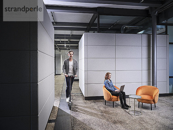 Geschäftsfrau sitzt im Sessel in einem modernen Büro mit Laptop und einem Kollegen auf einem Elektroroller