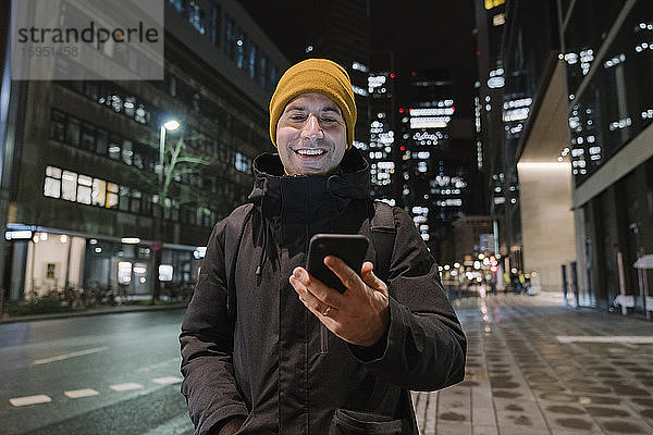 Porträt eines lächelnden Mannes  der nachts auf ein Smartphone schaut  Frankfurt  Deutschland