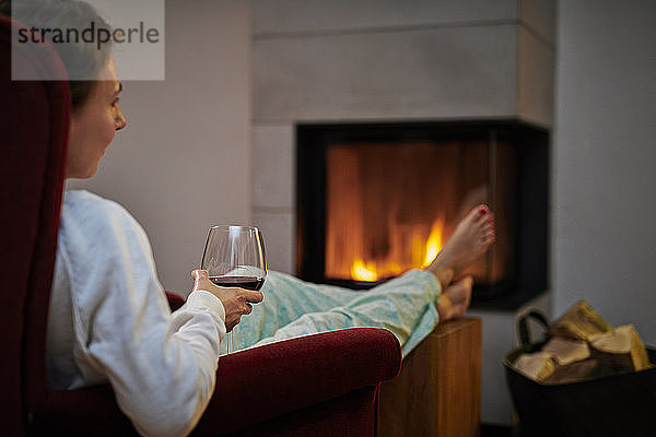 Frau sitzt auf einem Sessel mit einem Glas Rotwein und entspannt sich vor dem Kamin