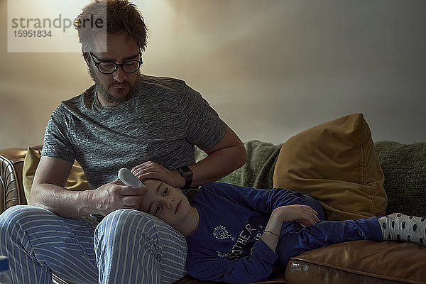 Mann hält Thermometer auf der Stirn der kranken Tochter im Wohnzimmer zu Hause