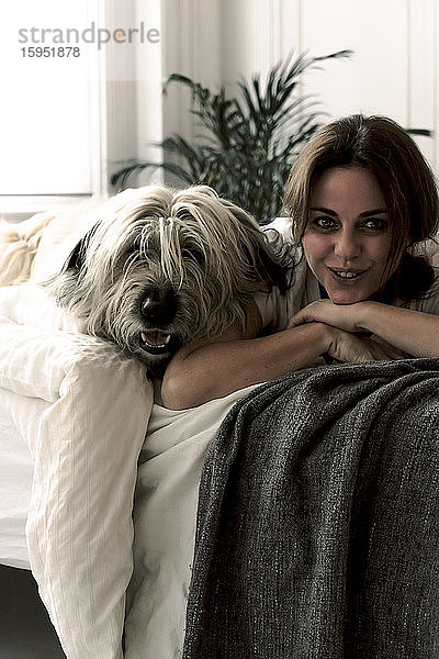 Porträt einer lächelnden  reifen Frau  die mit ihrem Hund auf dem Bett liegt