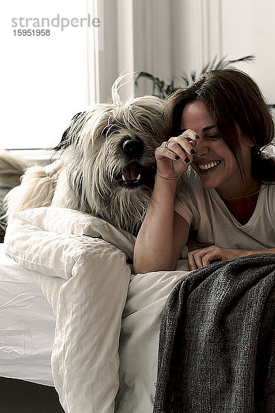 Lachende reife Frau liegt auf dem Bett und spielt mit ihrem Hund