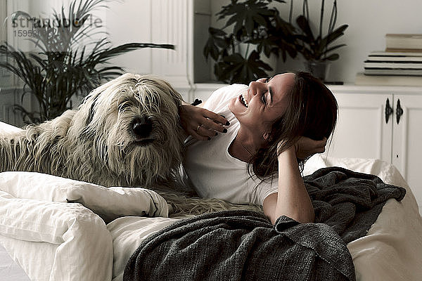 Lachende reife Frau liegt auf dem Bett und hat Spaß mit ihrem Hund
