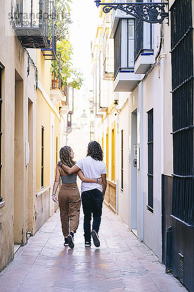 Rückansicht eines jungen Paares  das mit Armen auf einer engen Straße in Santa Cruz  Sevilla  Spanien  herumläuft