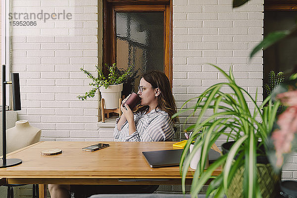 Nachdenkliche Frau trinkt Kaffee  während sie im Heimbüro am Schreibtisch sitzt