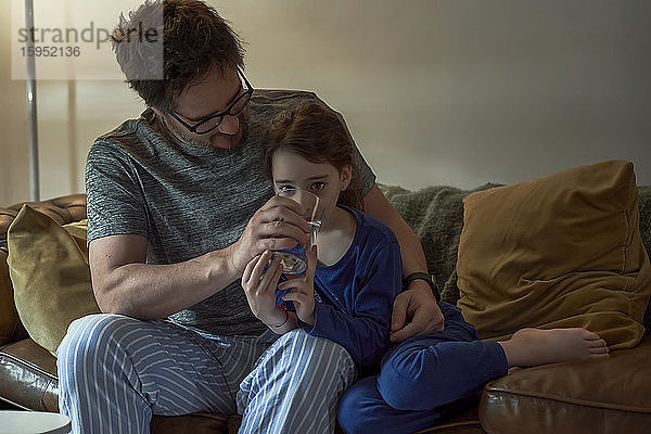 Vater hilft kranker Tochter im Trinkwasser  während er zu Hause auf dem Sofa im Wohnzimmer sitzt