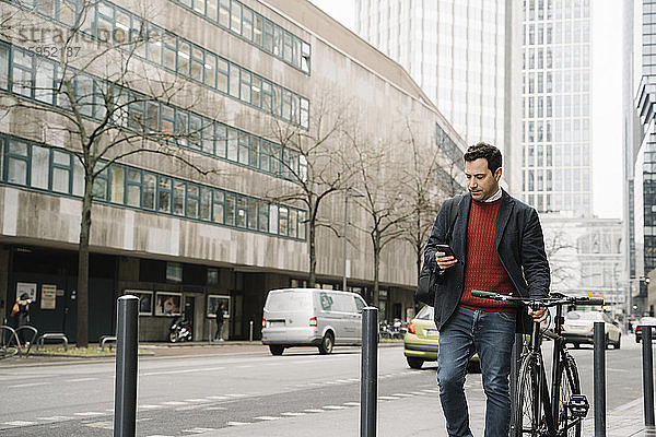 Geschäftsmann mit Fahrrad  der ein Smartphone benutzt  während er gegen Gebäude in der Stadt läuft  Frankfurt  Deutschland