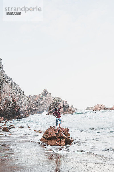 Junge Frau mit erhobenem Arm auf einem Felsen stehend  Praia da Ursa  Lissabon  Portugal